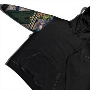 May include: Black hoodie with a front pocket and a hood. The left sleeve features a colorful, detailed tapestry design with green, blue, and gold tones. The hoodie is made of a soft, comfortable material.