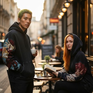 May include: Two people wearing black hoodies with tapestry sleeves are shown outdoors. One person holds a white cup, the other reads a book. The hoodies have a front pocket and a hood. The sleeves feature a colorful tapestry design.