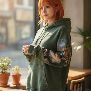 May include: An olive green hoodie with a front pocket and drawstring hood. The sleeves feature a detailed, colorful illustration. The person in the image is wearing the hoodie. The background includes a window, tables, and potted plants.