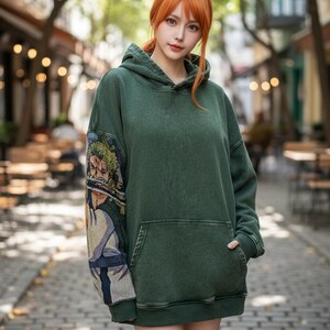 May include: A dark green hoodie with a front pocket and a hood. The left sleeve features a colorful tapestry design. The hoodie is worn in an outdoor setting with trees and a brick-paved walkway.
