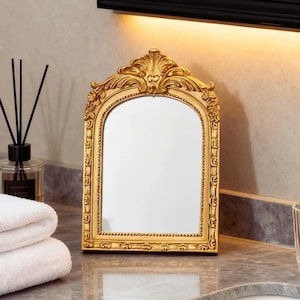 May include: A gold-colored, ornate framed mirror with an arched top. The frame features detailed scrollwork and floral designs. The mirror is placed on a countertop, with a stack of white towels and a diffuser in the background.