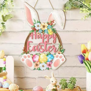 Puede incluir: Adorno de pared de madera con forma de conejito de Pascua con las palabras "Happy Easter" en escritura rosa. El diseño incluye orejas de conejo, huevos coloridos y detalles florales. Fondo de pared de ladrillo blanco.