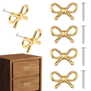 May include: Gold-colored bow-shaped cabinet knobs and pulls. The image shows several knobs with screws, and two installed on a wooden drawer. These decorative knobs add a touch of elegance to furniture.