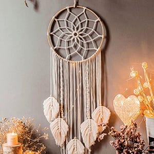 May include: A cream-colored macrame dream catcher with a circular wooden frame and intricate woven details. The dream catcher features hanging strands and decorative feather accents. It is a bohemian-style wall hanging.