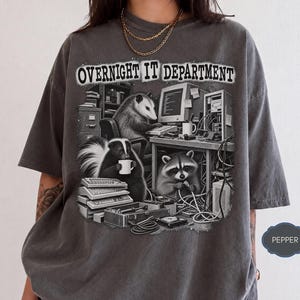 以下が含まれることがあります： OVERNIGHT IT DEPARTMENTというテキストがプリントされたチャコールグレーの半袖Tシャツ。イラストには、古いオフィス機器やケーブルが置かれたデスクで作業するオポッサム、スカンク、アライグマが描かれています。