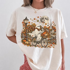 May include: An off-white t-shirt featuring a graphic of a ghost wearing a striped scarf and riding a bicycle through an autumn scene with falling orange leaves, a street lamp, pumpkins, a full moon, and a small village in the background.