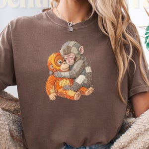 May include: Brown t-shirt with a graphic of two hugging monkeys. One monkey is orange and the other is grey. The shirt has the text "The Sentimental T". The shirt is worn with blue denim shorts and a cream-coloured sweater.