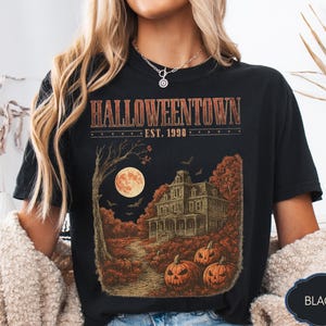 Könnte beinhalten: Ein schwarzes T-Shirt mit einem Grafikdruck und dem Text HALLOWEENTOWN EST. 1998. Das Design zeigt ein großes Haus, einen Vollmond, Fledermäuse und orangefarbene Kürbisse in einer Waldlandschaft. Das T-Shirt hat einen Rundhalsausschnitt.