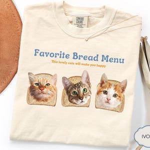 Könnte beinhalten: Cremefarbenes T-Shirt mit dem Text "Favorite Bread Menu" und drei Katzenköpfen in Toastscheiben. Der Text "This lovely cats will make you happy" steht darunter. Ein brauner Hut und dekorative Blätter sind im Hintergrund.