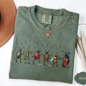 May include: A moss green t-shirt with a playful design of six insect musicians. The insects, including a beetle, grasshopper, and ladybug, are depicted playing instruments. A gold necklace is draped across the shirt.