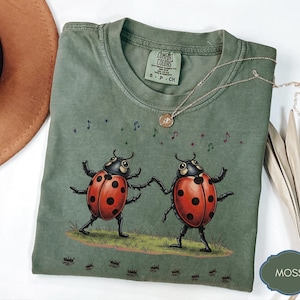 May include: A moss green short-sleeve t-shirt featuring a graphic of two red ladybirds with black spots dancing on a patch of grass. Musical notes and stars float above the ladybirds, while small black ants are printed along the bottom hem.