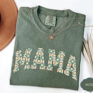 Op de afbeelding: Een mosgroen Comfort Colors T-shirt met het woord "MAMA" in een bloemenpatroon. Het shirt heeft een ronde hals en korte mouwen. Een gouden ketting met een hanger is over het shirt gedrapeerd.