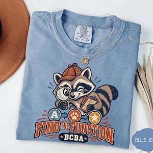 Könnte beinhalten: Ein hellblaues T-Shirt mit einer Grafik eines Waschbären, der einen Detektivhut trägt und eine Lupe sowie ein Klemmbrett hält. Unter dem Waschbären befinden sich die Buchstaben A, ein Pfotenabdruck und ein Stern sowie der Text FIND THE FUNCTION BCBA.