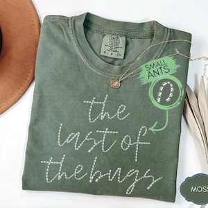 Könnte beinhalten: Ein moosgrünes Kurzarm-T-Shirt mit dem in Weiß gestickten Text "the last of the bugs" auf der Vorderseite. Das Shirt verfügt über ein Comfort Colors Markenetikett am inneren Halsausschnitt und eine kleine kreisförmige Grafik mit "SMALL ANTS".