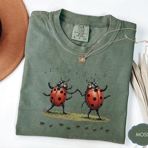 May include: A moss green t-shirt featuring a graphic of two red ladybugs with black spots dancing on a patch of grass. Small musical notes float above the ladybugs, and a line of tiny ants marches below them. The shirt has a Comfort Colors brand tag.
