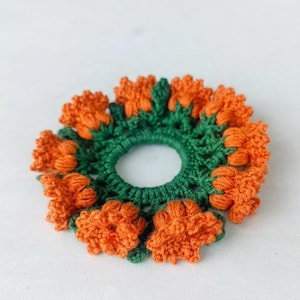 May include: A handmade crochet hair tie with a floral design. The hair accessory has a green base with orange flower petals. The circular design is suitable for various hairstyles.