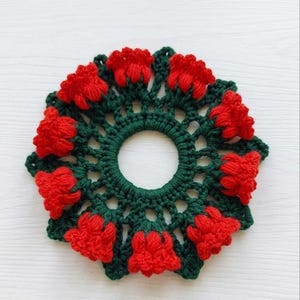 May include: A handmade crochet coaster featuring a floral design. The coaster has a dark green base with a ring of red flowers around the edge. The coaster is circular with a hole in the center.