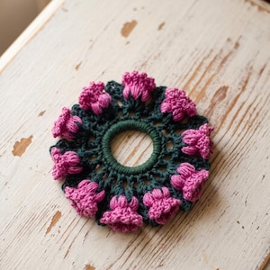 May include: A handmade crochet hair tie featuring a dark green base with pink flower accents. The hair accessory is circular, with a green elastic band in the center. The flowers are evenly spaced around the edge of the hair tie.