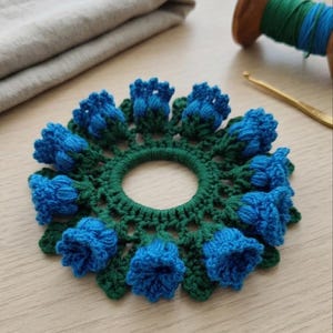 Könnte beinhalten: Ein handgefertigter, blau-grüner Häkeluntersetzer mit floralem Design. Der Untersetzer hat einen zentralen Ring, der von blauen Blütenknospen und grünen Blättern umgeben ist. Der Untersetzer liegt auf einer Holzoberfläche.