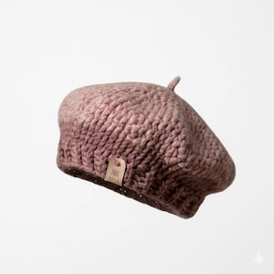 Op de afbeelding: Een stoffige roze baret van dik gebreide wol. De baret heeft een afgeronde kroon en een kleine, omhoog wijzende steel aan de bovenkant. Een klein, rechthoekig label met de tekst "DURAS WASA" is bevestigd. Een modieus accessoire.