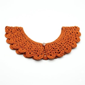 May include: An orange crochet collar with a scalloped edge and a single button closure. The collar is made of a textured yarn and is designed to be worn around the neck. The collar is a warm, earthy tone.