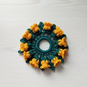 Könnte beinhalten: Ein handgefertigtes Häkel-Haargummi mit floralem Design. Das Haaraccessoire hat eine tealfarbene Basis mit gelben Blumenakzenten. Das kreisförmige Design ist mit einer tealfarbenen Öffnung zentriert. Das Haargummi ist aus Garn gefertigt.
