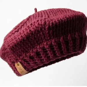 Op de afbeelding: Bordeauxrode gebreide baret met een kabelgebreid patroon. De baret heeft een kleine, ronde bovenkant en een leren label aan de zijkant. De hoed is gemaakt van een dik, warm materiaal.