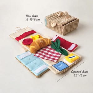 Könnte beinhalten: Ein beiger Picknickkorb aus Stoff mit Griff, begleitet von einer Reihe von Plüsch-Lebensmitteln. Enthält ein Croissant, rot-weiß kariertes Tuch und ein blaues Tuch mit weißen Klettverschlüssen. Die Boxgröße beträgt 16x15x8 cm, und die geöffnete Größe beträgt 28x43 cm.