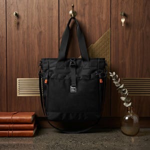 May include: A black tote bag featuring a large front mesh pocket, a central buckle strap, and two top handles. The bag includes side drawstrings with orange toggles and a small rectangular label with the text Spurn centered on the mesh pocket.