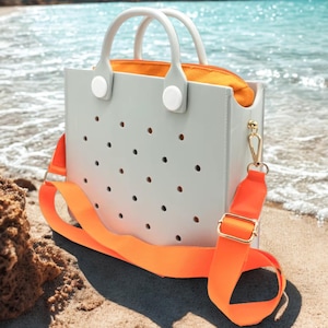 May include: A pale grey tote bag with a perforated pattern, white handles, and an orange interior. It has an orange shoulder strap with gold-coloured hardware. The bag is on a sandy beach with the sea in the background.