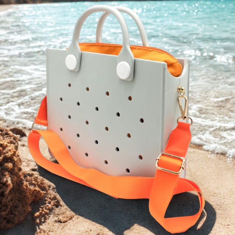 May include: A pale grey tote bag with a perforated pattern, white handles, and an orange interior. It has an orange shoulder strap with gold-coloured hardware. The bag is on a sandy beach with the sea in the background.