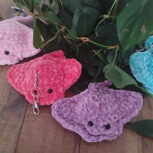 May include: A collection of handmade, plush stingray keychains in various colors: pink, red, purple, and blue. Each keychain features black button eyes and a silver-colored clasp. The keychains are arranged on a wooden surface with green foliage.