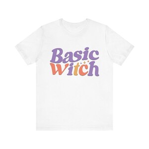 May include: A white short-sleeve t-shirt featuring the text Basic Witch printed in a stylized, wavy font. The word Basic is in purple, and the word Witch is in a mix of orange and purple, accented with small four-pointed star icons.