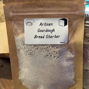 Puede incluir: Una bolsa de papel marrón llena de un polvo gris claro, etiquetada como "Artisan Sourdough Bread Starter". La etiqueta incluye ilustraciones de pan. La bolsa sellada contiene 30 gramos de masa madre.