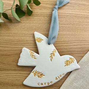 May include: White ceramic dove ornament with gold leaf detailing and the phrase "carpe diem." The dove is suspended by a light blue ribbon. Eucalyptus leaves are visible in the background. Decorative item.