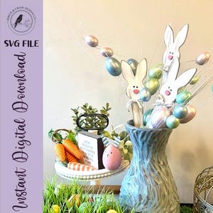 May include: An Easter-themed display featuring a textured blue vase with wooden bunny cutouts and pastel-colored eggs on wire stems. A white tiered tray holds carrots, a pink egg, and a small sign. The words "SVG File" and "Instant Digital Download" are visible.