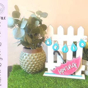 Puede incluir: Decoración de primavera con una valla blanca, un paraguas rosa con la palabra "spring" y una pancarta con "hello" en forma de gotas de lluvia azules. Una planta verde en una maceta texturizada está a la izquierda.