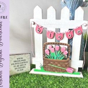 May include: A wooden spring-themed display with a white picket fence and a "BLOOM" banner. A basket of pink tulips is placed in front of the fence. A small sign reads "YOU'RE PRETTY MUCH MY MOST FAVORITE OF ALL TIME IN THE HISTORY OF EVER". The display is set on a green surface.
