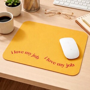 Puede incluir: Alfombrilla de ratón de color amarillo mostaza con la frase "i love my job" en rojo. Un ratón de ordenador blanco descansa sobre la alfombrilla. La alfombrilla está sobre un escritorio de madera clara, con un teclado, gafas y un bloc de notas en el fondo.