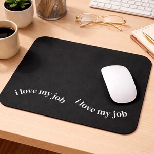 Puede incluir: Alfombrilla de ratón rectangular negra con el texto "i love my job" en blanco. Un ratón de ordenador blanco está en el lado derecho de la alfombrilla. La alfombrilla está sobre un escritorio de madera marrón claro.