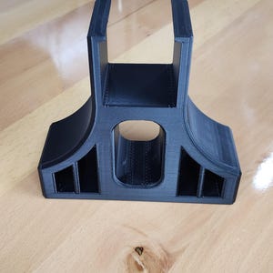 May include: A black 3D-printed stand with a unique design. It features a rectangular base with an arched top and two vertical supports. The stand has an oval opening in the center and rectangular cutouts on the sides.