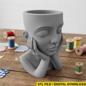Op de afbeelding: Een grijze, gebeeldhouwde kopvormige plantenbak met gesloten ogen, rustend op handen. De plantenbak staat op een houten oppervlak met spoelen garen in verschillende kleuren. De tekst "STL FILE • DIGITAL DOWNLOAD" staat onderaan.