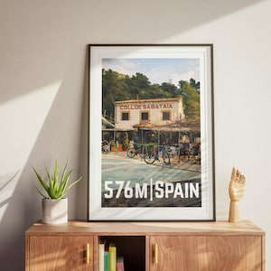 May include: Framed art print depicting a Spanish cafe with bicycles, with the text "COLL DE SABATAIA" above and "576M SPAIN" below. The print is displayed on a wooden cabinet. The height is 576 metres.