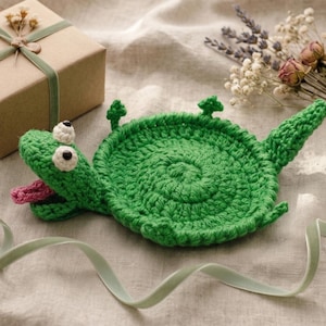 Puede incluir: Un posavasos de ganchillo verde hecho a mano con forma de tortuga, con ojos blancos y una lengua rosa. El posavasos está sobre una superficie beige con una caja de regalo envuelta y flores secas en el fondo.