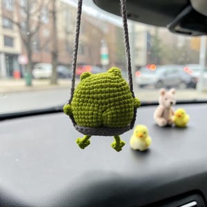 May include: A handmade, green crochet frog ornament swinging from a gray string. The frog is suspended in front of a car dashboard, with a small teddy bear and two yellow ducklings in the background.