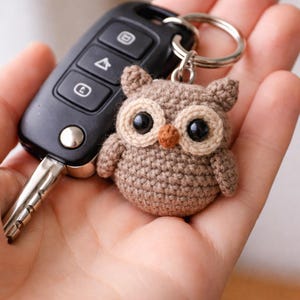 Może przedstawiać: Szydełkowy brelok do kluczy w kształcie sowy w odcieniach brązu, beżu i czerni. Sowa ma duże oczy i mały, pomarańczowy dziób. Brelok jest przymocowany do srebrnego kółka, obok pilota do samochodu.