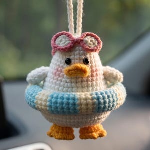 May include: A handmade crochet duck ornament with a white body, pink cheeks, and an orange beak and feet. The duck wears pink sunglasses and a blue and white striped inner tube. A white string is attached for hanging.