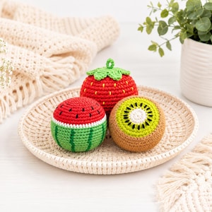 May include: Three crocheted fruit-shaped items are presented on a woven plate. A red strawberry with green leaves, a green and red watermelon, and a brown and green kiwi are displayed. The plate is on a white surface, with a cream-coloured crocheted blanket in the background.