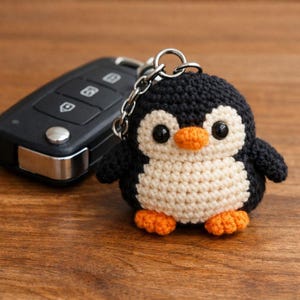 May include: A handmade crochet penguin keychain. The penguin has a black body, white belly, and orange feet and beak. It is attached to a silver chain and keyring. A black car key fob is in the background.