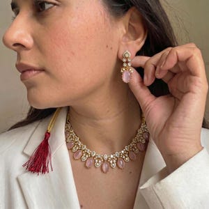 May include: A gold-toned necklace and earring set featuring pink oval-shaped gemstones and clear crystals. The necklace has a tassel and the earrings are dangling. The jewelry is worn with a white blazer.